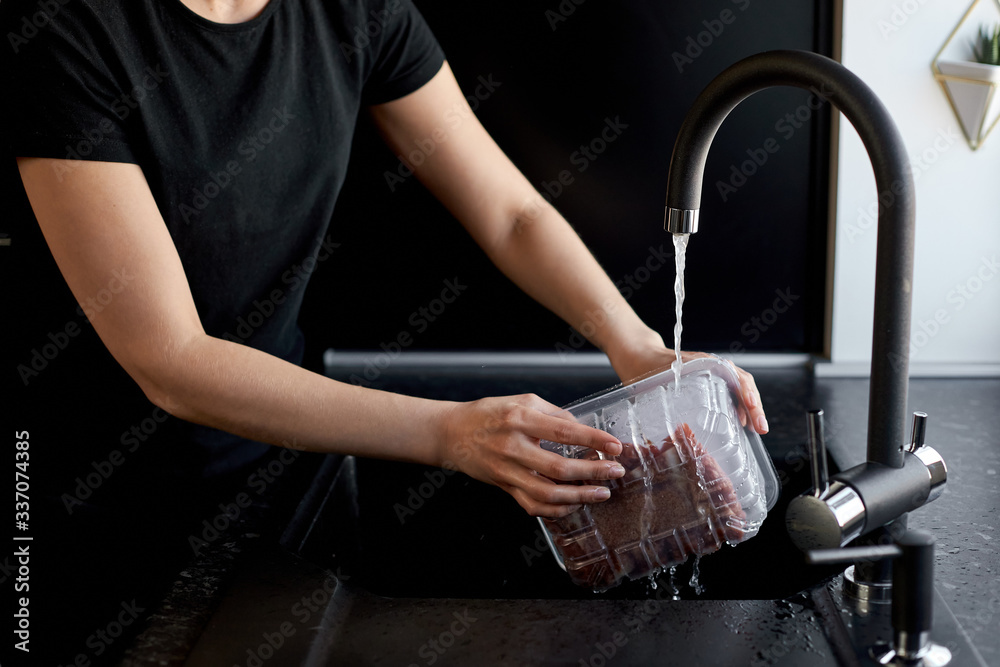 woman washes a package of meat under water in a black kitchen