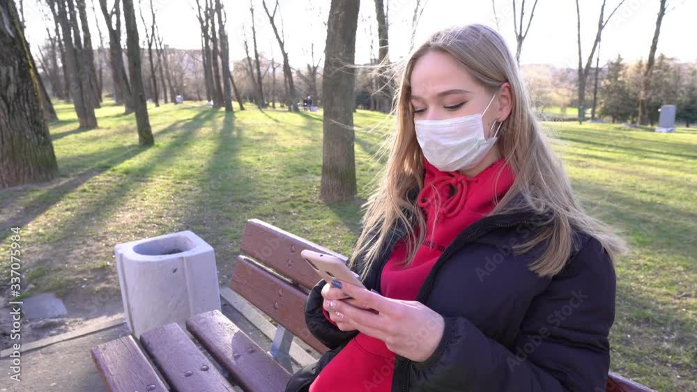 Young woman in red in protective mask using smartphone for calling or recording video in the city park. Pandemic Covid-19 coronavirus protection. I sat down to rest on a bench by the river. 4k