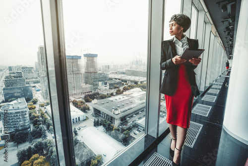 A good-looking African-American woman entrepreneur in a red skirt and black jacket is using a digital tablet while leaning against a panoramic window of a business office high-rise, cityscape outside