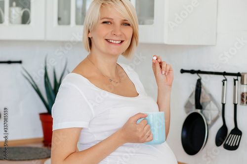 A young blonde pregnant girl in a white T-shirt and gray pants stands in the kitchen with a thermometer and pills in her hands
