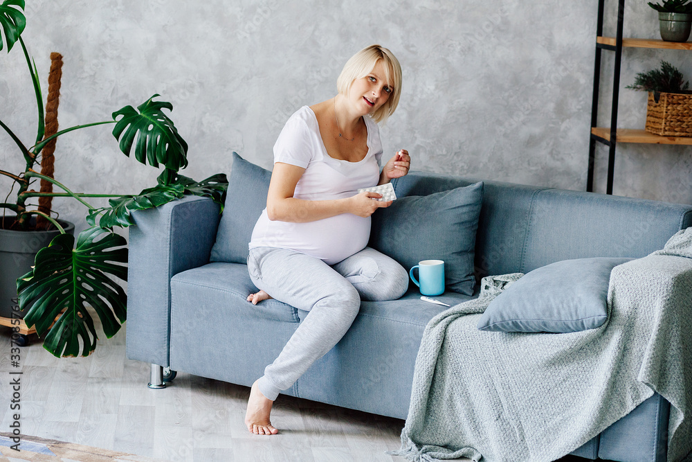 A young pregnant blonde in a white T-shirt and gray trousers sits on a gray sofa and takes pills, vitamins A young pregnant blonde in a white T-shirt and gray trousers sits on a gray sofa and takes pills, vitamins