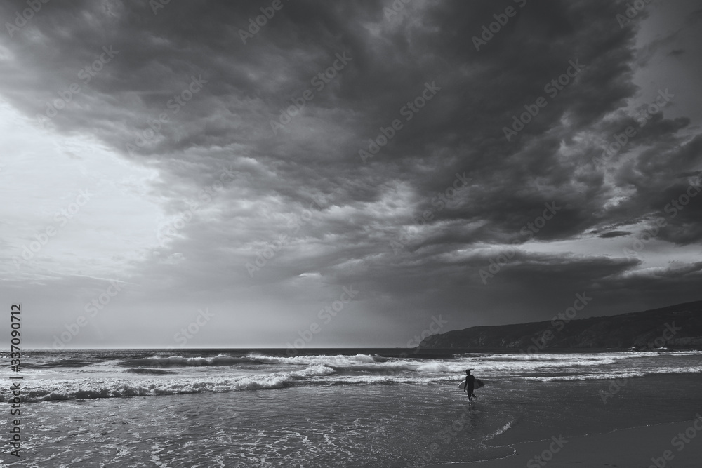 Lone surfer walks into the ocean to catch a wave. View on an Atlantic coast and dramatic cloudy sky at sunset. Concept of outdoor activities. Lisbon, Portugal.High Contrast. B&W color.