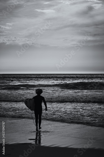 Lone surfer walks into the ocean to catch a wave. View on an Atlantic coast and dramatic cloudy sky at sunset. Concept of outdoor activities. Lisbon, Portugal.High Contrast. B&W color.