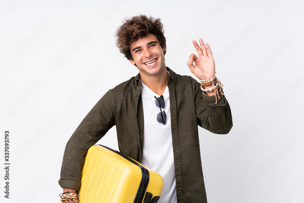 Young Traveler man holding a suitcase over isolated white background showing an ok sign with fingers Young Traveler man holding a suitcase over isolated white background showing an ok sign with fingers