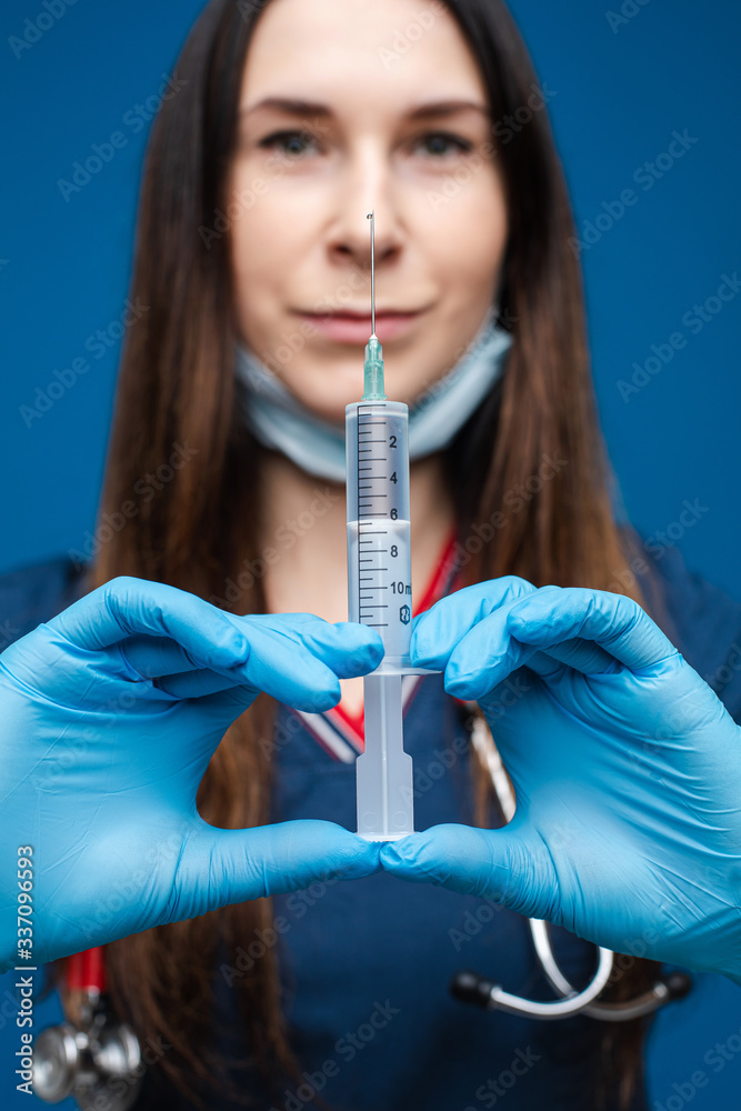 Portrait of caucasian female with ling dark hair in medical gown, blue medical gloves and special mask holds a syringe with medicine