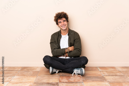 Young caucasian man sitting on the floor with arms crossed and happy