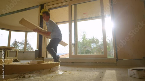 SLOW MOTION LENS FLARE, LOW ANGLE: Young Caucasian man wearing a yellow hard hat and carrying a long plank trips while entering the glued-laminated timber house under construction. Clumsy builder.