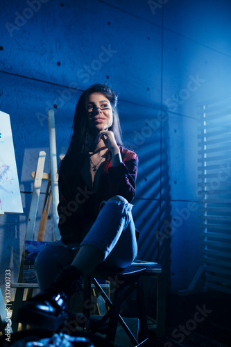 The artist in a plaid shirt sitting in a dark smoky room Studio near the easels and looking forward.