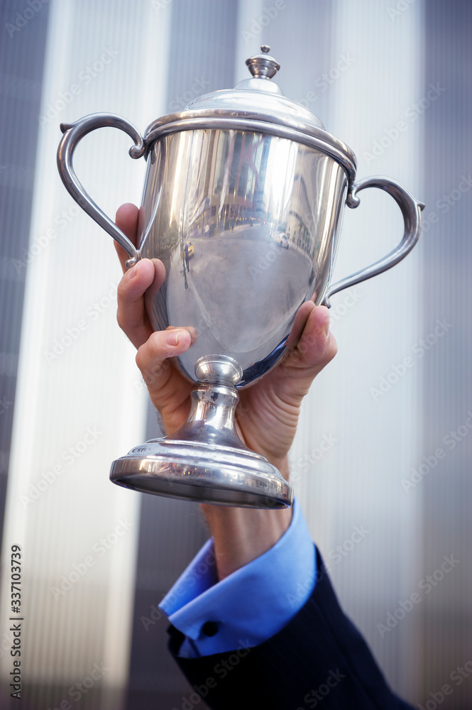 Fototapeta premium Hand of proud unrecognizable businessman holding up a silver trophy outdoors in front of shiny modern office building