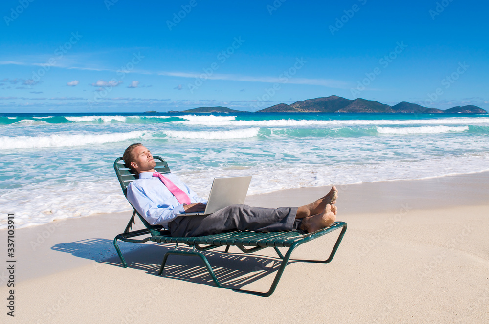 Barefoot businessman working on his laptop on a beach chair relaxing on the shore of an empty tropical beach