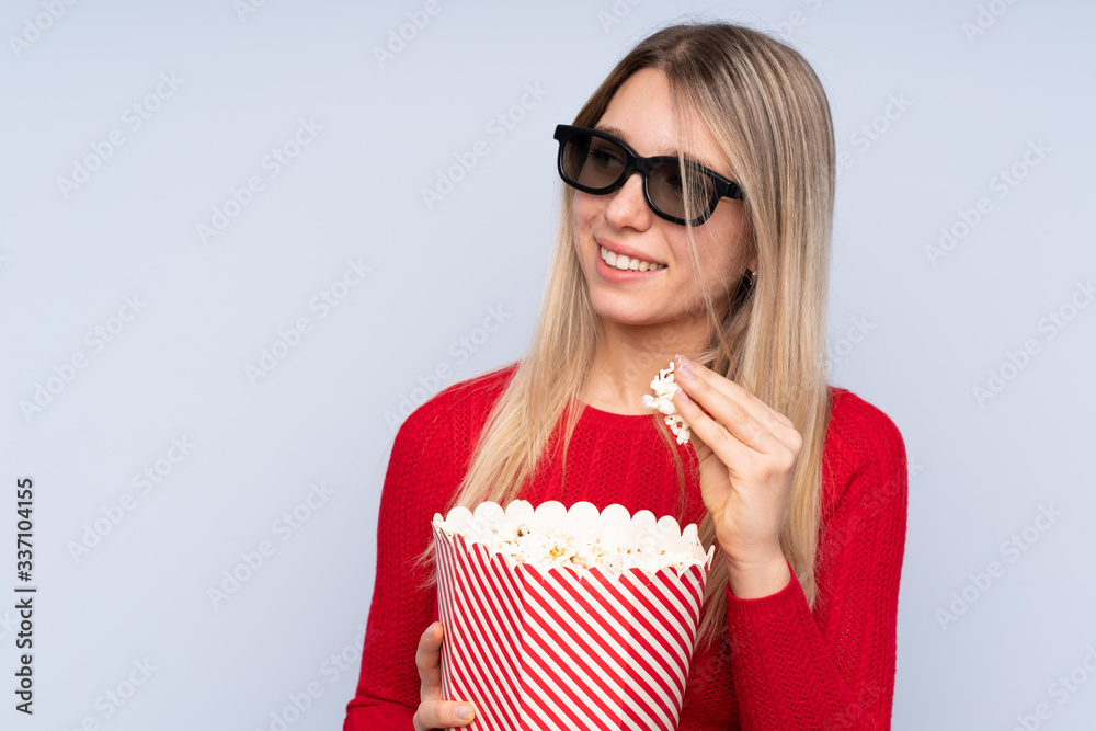Young blonde woman over isolated blue background with 3d glasses and holding a big bucket of popcorns