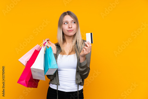 Young blonde woman over isolated yellow background holding shopping bags and a credit card and thinking