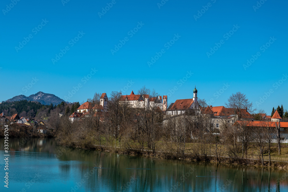 Obraz premium Die Altstadt von Füssen auf der anderen Seite des Lechs mit Hohem Schloss, St. Mang mit Kloster und Kirche und der Spitalkirche bei blauem Himmel