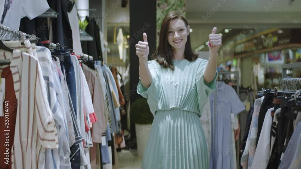 Positive brunette woman is giving thumbs up with both hands and looking at the camera while standing inside the shop. Front view of cheerful beautiful woman showing thumbs up at the camera in the Positive brunette woman is giving thumbs up with both hands and looking at the camera while standing inside the shop. Front view of cheerful beautiful woman showing thumbs up at the camera in the