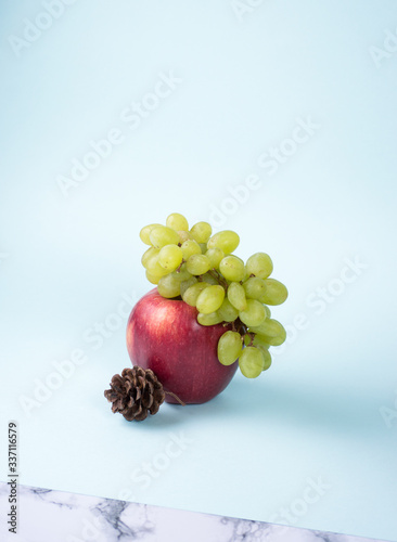 Branch of green grape lies on big red apple wih pine cone on blue background