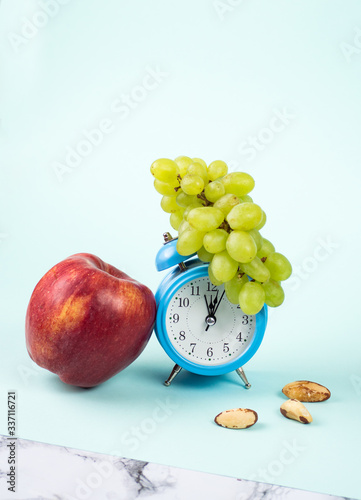 Bright blue alarm clock with branch of green grape on it on blue background with big red apple and nuts