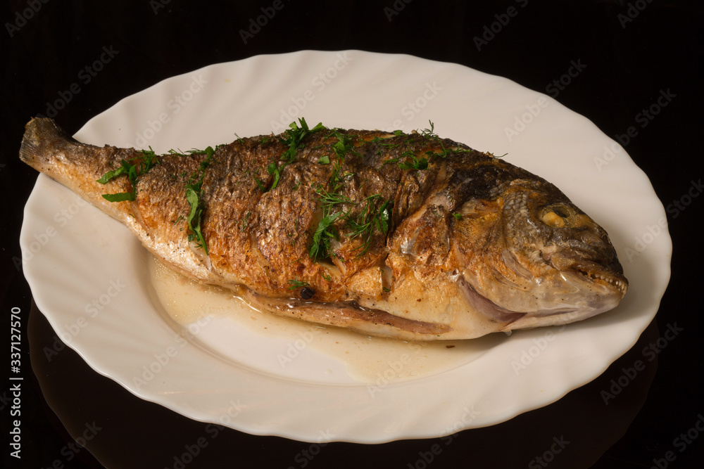 grilled whole fish with seasonings on a white plate. On the background of a reflective surface grilled whole fish with seasonings on a white plate. On the background of a reflective surface