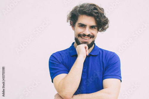 Positive happy guy leaning chin on hand, looking at camera, smiling. Handsome bearded young man in blue casual t-shirt posing isolated over white background. Male portrait concept