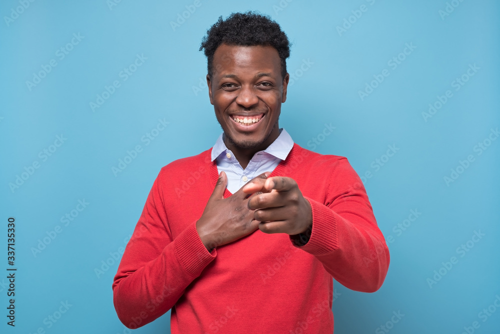 Young handsome african american man laughing pointing at you on joke, isolated on blue wall background. Positive human emotion facial expression feelings Young handsome african american man laughing pointing at you on joke, isolated on blue wall background. Positive human emotion facial expression feelings