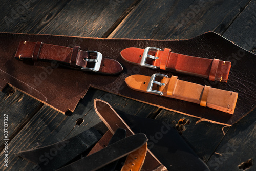 Handmade brown tone watch straps with steel buckle laying on wooden rustic surface next to leather cuttings and leather thread. Different leather colour tones.