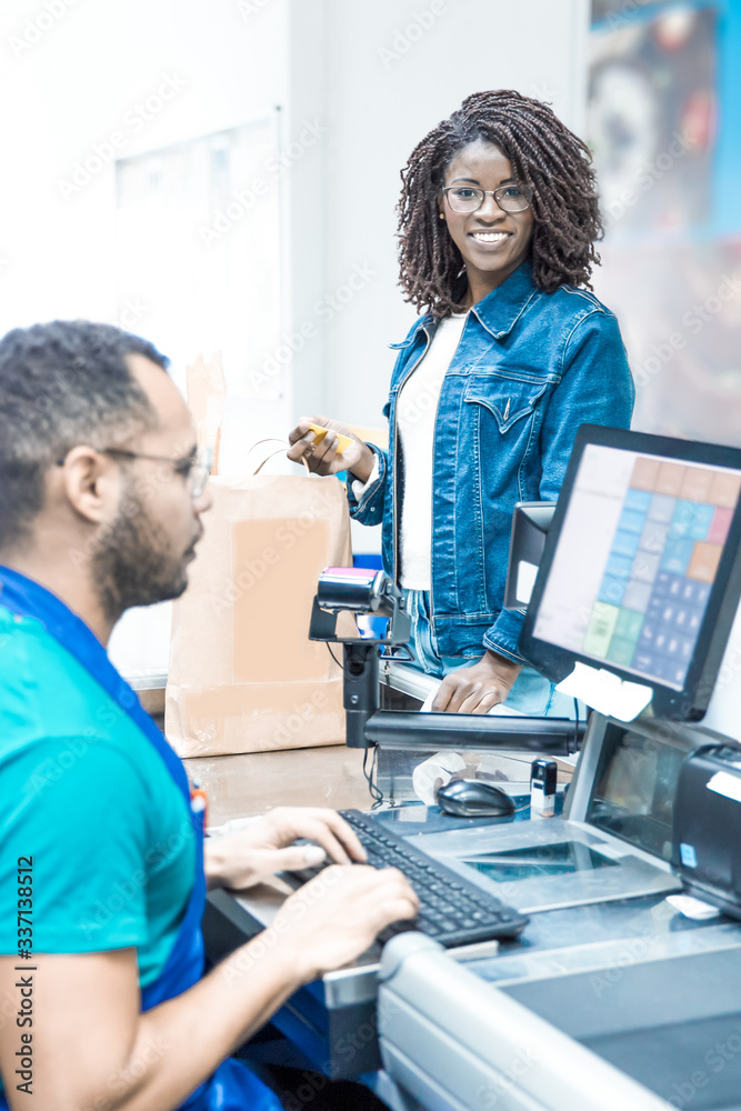 Obraz premium Smiling woman standing with credit card and packed food. Male cashier typing on keyboard at workplace. Shopping concept