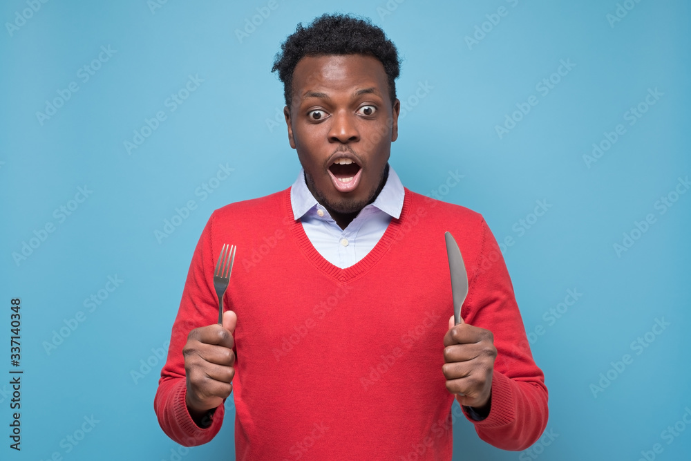 Fotografija hungry young african american man with a knife and a fork is shocked with tasty meal