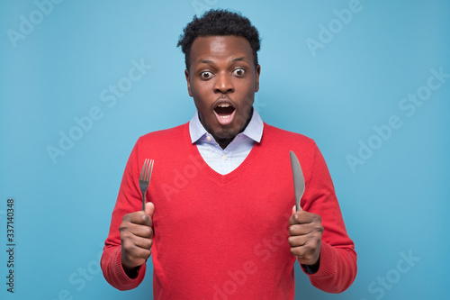 Fototapeta hungry young african american man with a knife and a fork is shocked with tasty meal