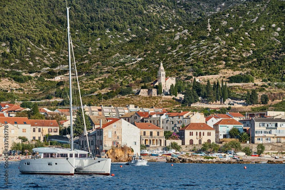 Seascape of city Komiza - the one of numerous port towns in Croatia, Catamaran in the foreground, orange roofs of houses, a cathedral St.Nicholas, Picturesque slopes of mountains