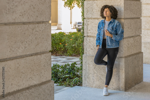 The slender fashionably modern woman relaxes by leaning against a stone wall. She has her eye on something going on in the distance, with one leg up on the wall holding her denim jacket. photo