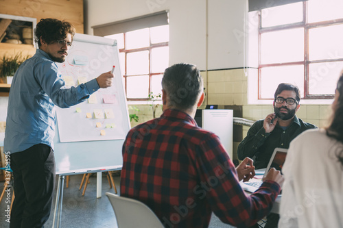 Creative team discussing ideas for startup project, speaker at board pointing at audience. Business colleagues in casual working together in contemporary office space. Brainstorming concept