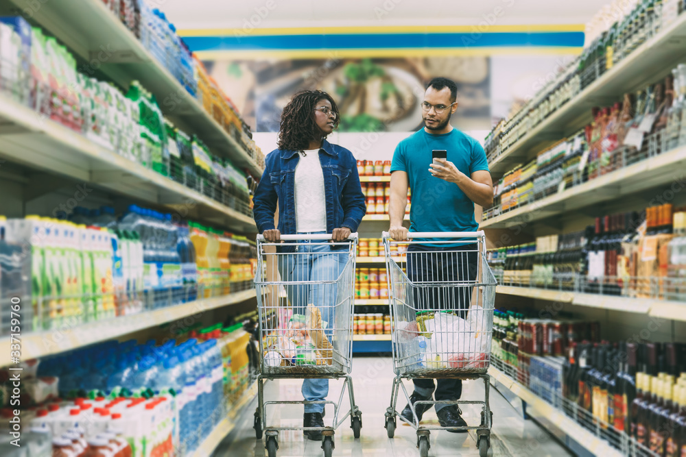 Peaceful young couple making purchases in grocery store. Bearded man in eyeglasses holding smartphone. Young woman with dreadlocks looking at boyfriend. Shopping concept