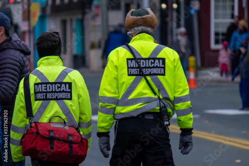 Two medical first responders walking in a street with people in the background. The officers are wearing a bright yellow reflective coat with grey stripes and a red first aid bag.  © Dolores  Harvey
