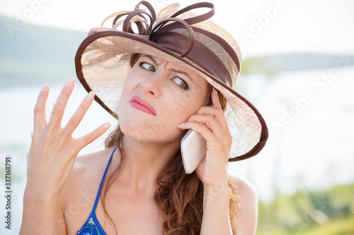 Portrait ot beautiful angry looking woman in hat raising hand frustrated talking on her mobile phone with gestures outside the city, lake sea background.  Negative human emotion, face expression photo