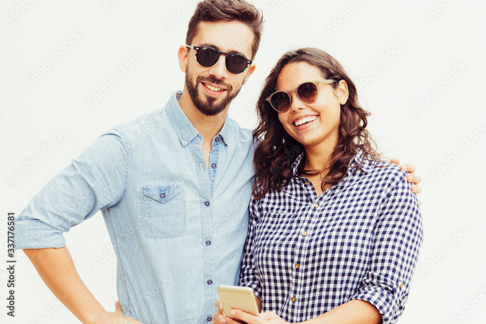 Obraz premium Positive couple of tourists wearing sunglasses, using smartphone, smiling at camera. Young woman in casual and man in glasses in glasses posing isolated over white background. Vacation concept