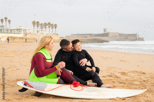 Happy family sitting on sand near surfboard. Father holding his son. Boy laughing. Mother looking aside. People in full body swimsuits. Vacation, surfing and summer concept