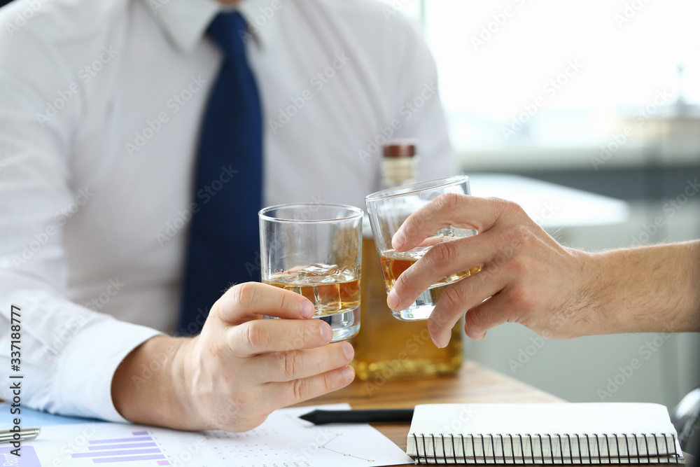 Close-up of office workers clink glasses after making good deal. Partners celebrating signing important agreement with cognac. Papers and documents on table. Business concept Close-up of office workers clink glasses after making good deal. Partners celebrating signing important agreement with cognac. Papers and documents on table. Business concept
