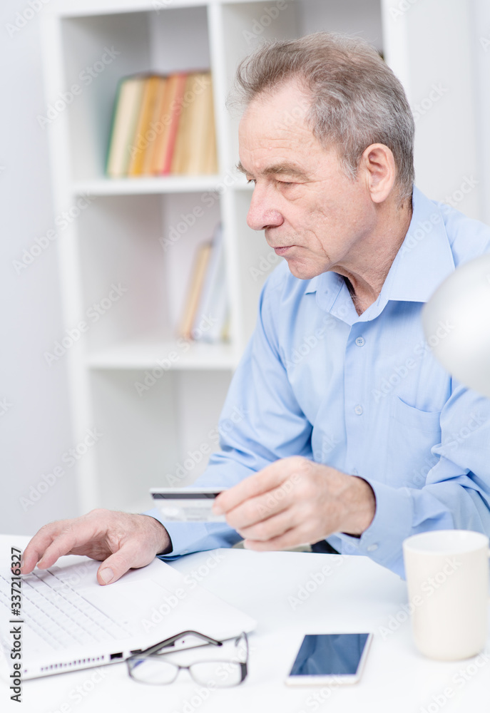 Senior citizen man working on a laptop holding a credit card in his hand looking at the screen of his computer Senior citizen man working on a laptop holding a credit card in his hand looking at the screen of his computer