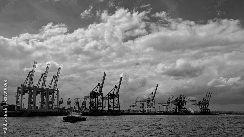 Obraz premium Panoramic view of the Port of Hamburg with cranes on the background along the river and a boat on water, under a cloudy sky, in black and white.