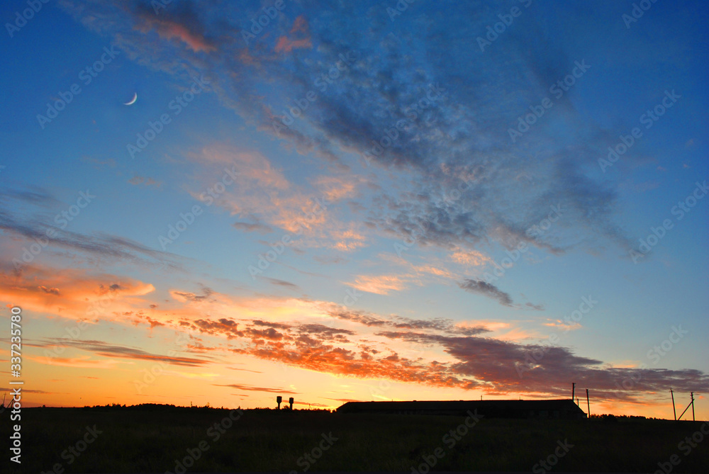 Beautiful sunset with moon over farm silhouette, sunlight on horizon of golden pink sky with clouds