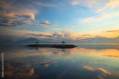 Sunrise seascape. Mountains and Agung volcano. Traditional gazebos on an artificial island in the ocean. Water reflection. Sanur beach, Bali, Indonesia.