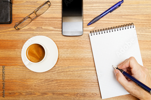 Wooden desktop with blank notebook, eyeglasses, cup of coffee and smartphone. Human hand with a pen writing in a notebook. Top view. Copyspace.