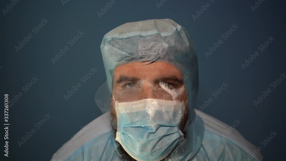 A tired bearded doctor breathes in a foggy mask and looks at the camera. Close-up. Sweat flows down the medical mask while breathing. A tired bearded doctor breathes in a foggy mask and looks at the camera. Close-up. Sweat flows down the medical mask while breathing.