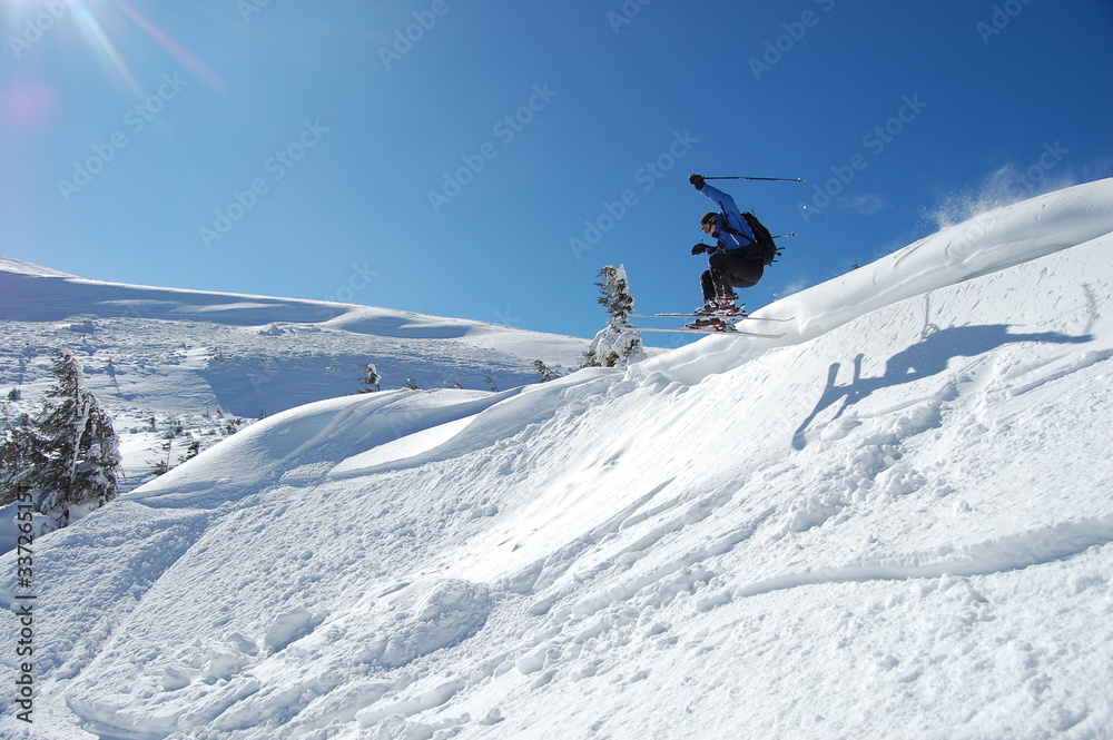 Skier in action on a sunny winter day.  High jump from the hill followed by snow powder. Blue sky and white snowy mountains on background. Extreme winter sports.