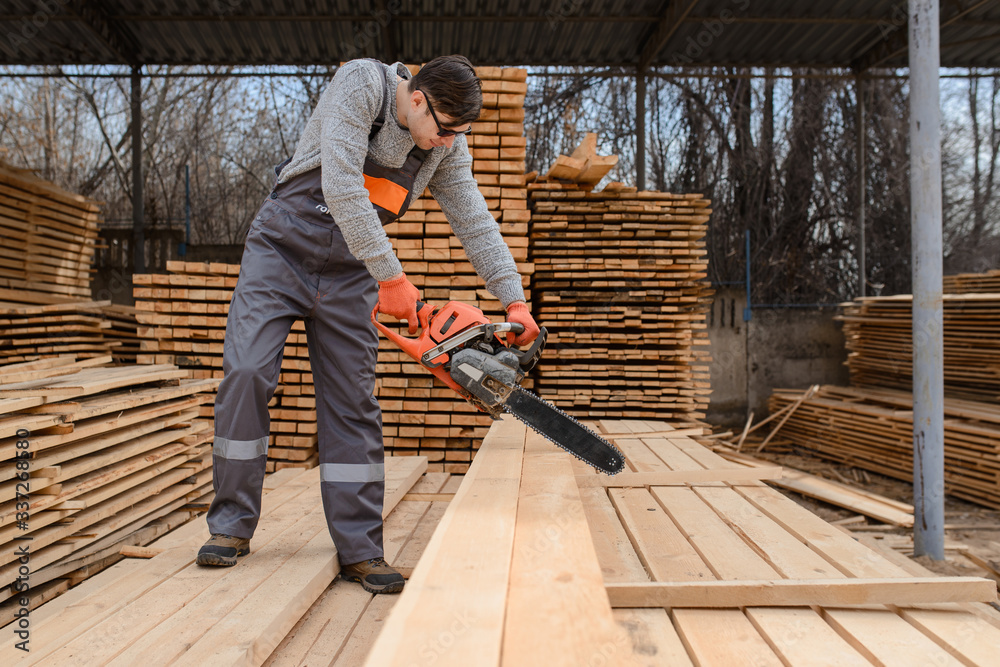 A portrait of young man carpenter at the wooden warehouse hold in his hands a chainsaw A portrait of young man carpenter at the wooden warehouse hold in his hands a chainsaw