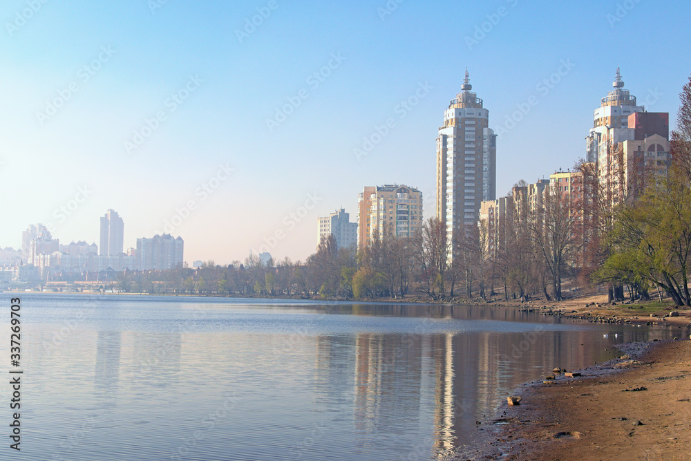 Obraz premium Scenic spring landscape view of Dnieper River and empty beach in modern Obolon district. Embankment with skyscrapers in morning smog in the background. Buildings reflected in the water. Kyiv, Ukraine