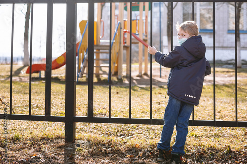 A boy in a medical mask stands next to a metal fence. A child yearns for playing on the Playground during quarantine