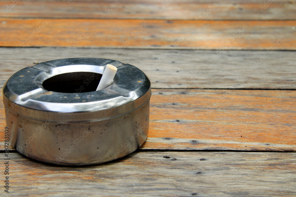 Round shape stainless ashtray is on light brown wooden floor and a cigarette in it, Thailand. Round shape stainless ashtray is on light brown wooden floor and a cigarette in it, Thailand.
