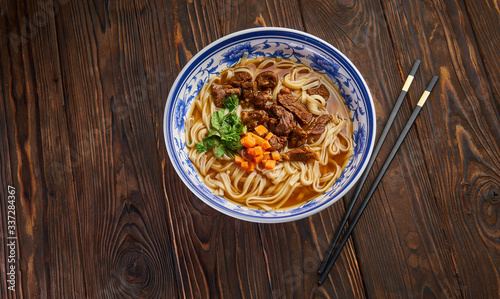 Chinese beef noodle soup in traditional blue bowl, fresh herbs and sliced carrot, pair of chopsticks on dark wooden background and copy space. Asian food concept top view