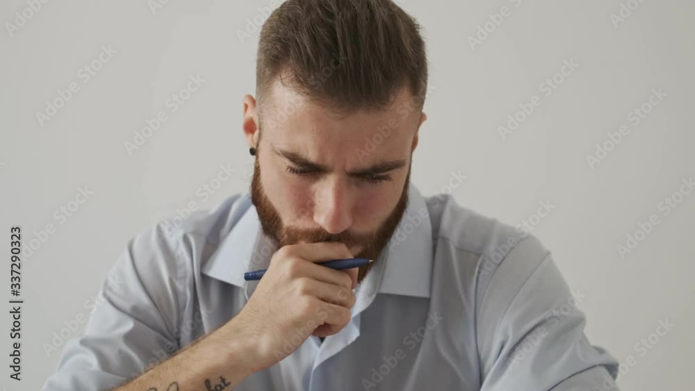 A concentrated serious young bearded man wearing a shirt is working sitting at the table over a white wall A concentrated serious young bearded man wearing a shirt is working sitting at the table over a white wall