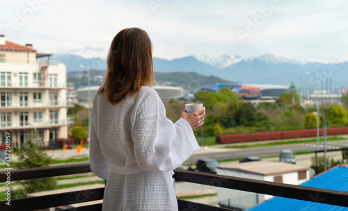Fototapeta Naklejka Na Ścianę i Meble -  Woman in a bathrobe on a balcony drinks coffee and looks at the street